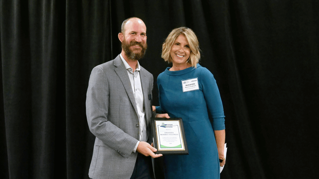 A man in a button-up shirt and jacket hands a plaque to a woman in a teal dress against a black backdrop
