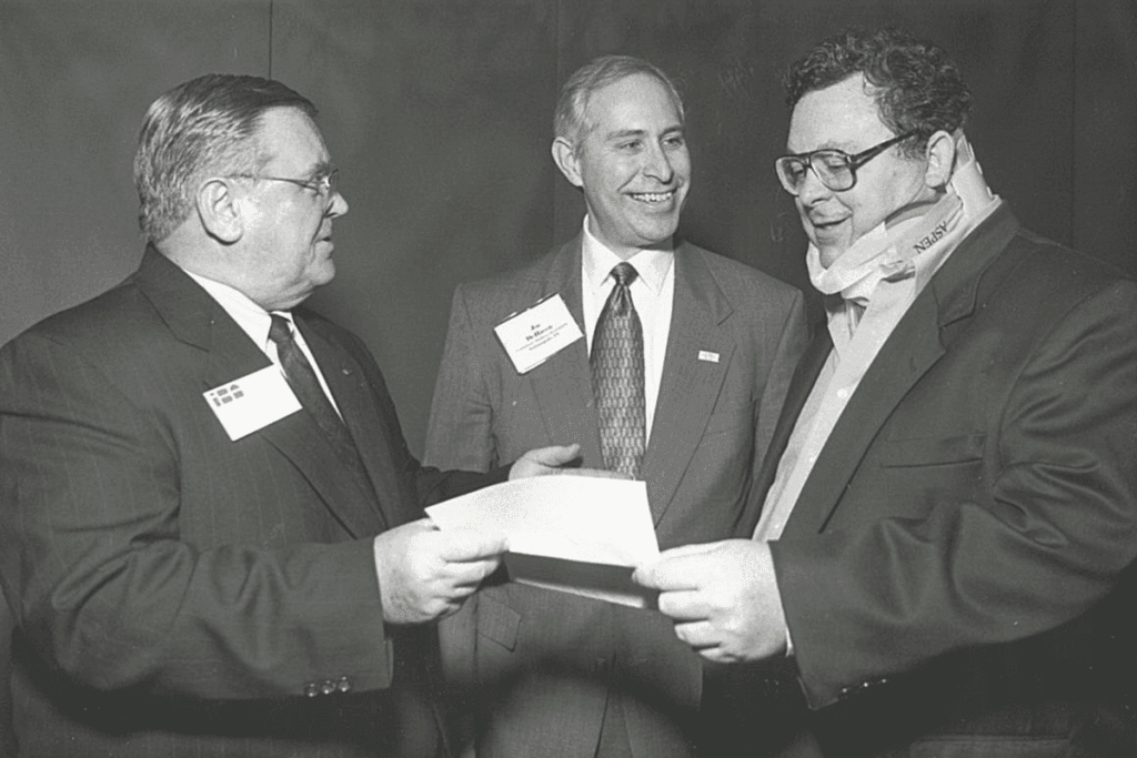 Three men in suits and ties stand in a circle and smiling as they discuss the contents of a paper being held in front of them