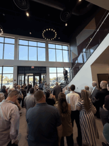 A group of people stand inside a building's lobby looking on as a man in a suit speaks from a staircase.
