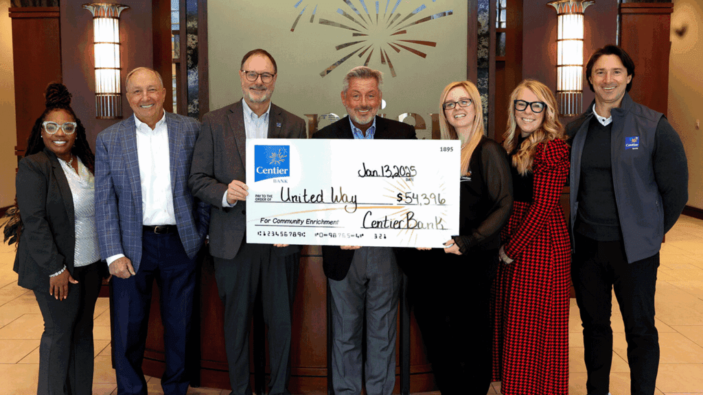 a photo showing seven people in professional clothing inside a bank building holding a ceremonial large check depicting a charitable donation