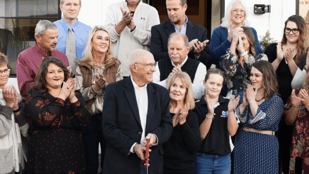a photo showing more than a dozen people in business clothing applauding after the ceremonial ribbon cutting outside a new bank facility