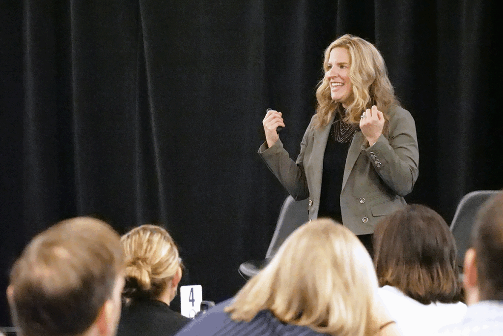 a woman wearing a dark top and lighter jacket speaks from the stage with a black backdrop at a conference