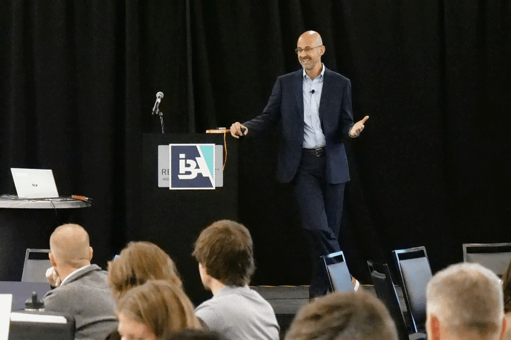 a man in a dark suit and light button-down shirt leans against a podium with the IBA logo on it as he speaks from the stage with a black backdrop at a conference