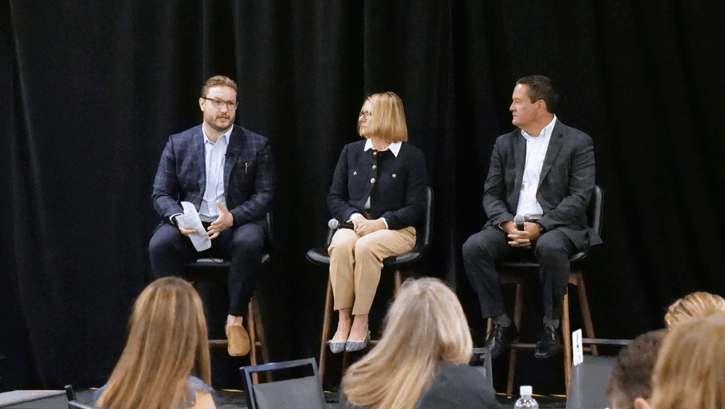 a man in a suit leads a panel discussion with another man and woman in professional clothing, all sitting on stools, on a stage with a black backdrop