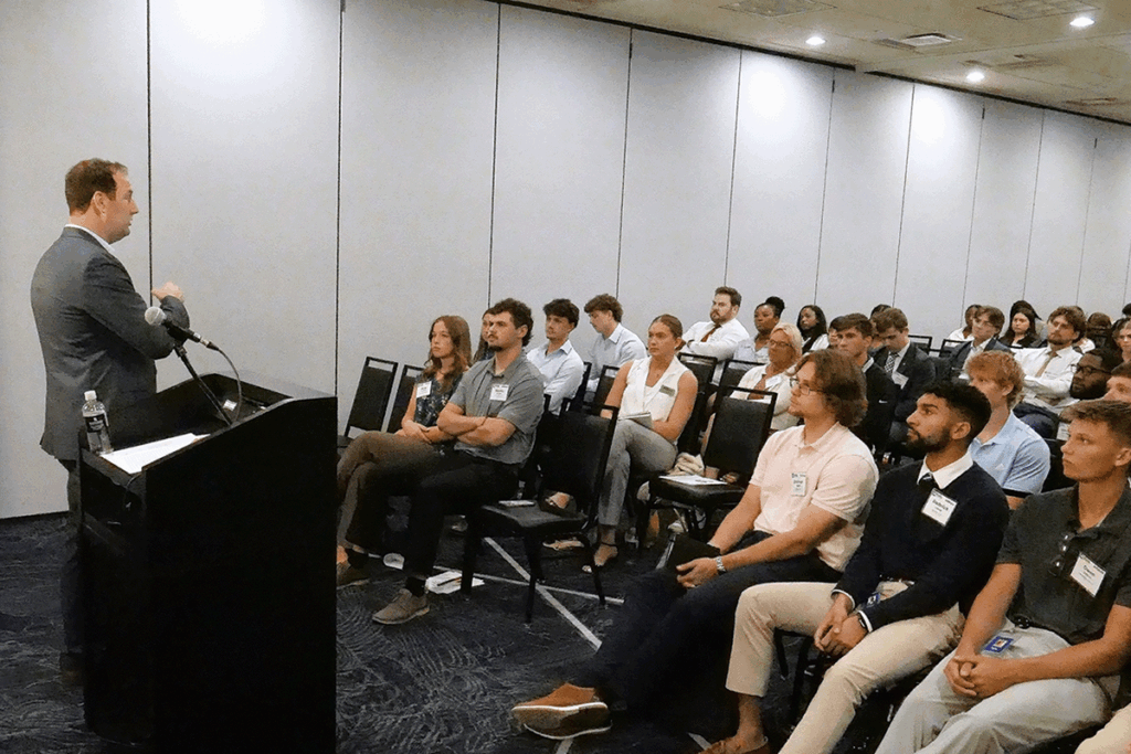 dozens of college students sit in chairs listening to a man speak from next to a podium