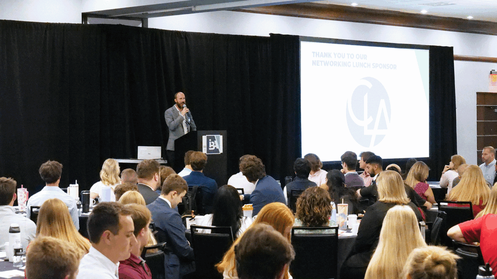 a crowd of attendees looks on as the host of a conferences addresses the crowd from a stage with black curtains behind him
