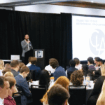 a crowd of attendees looks on as the host of a conferences addresses the crowd from a stage with black curtains behind him