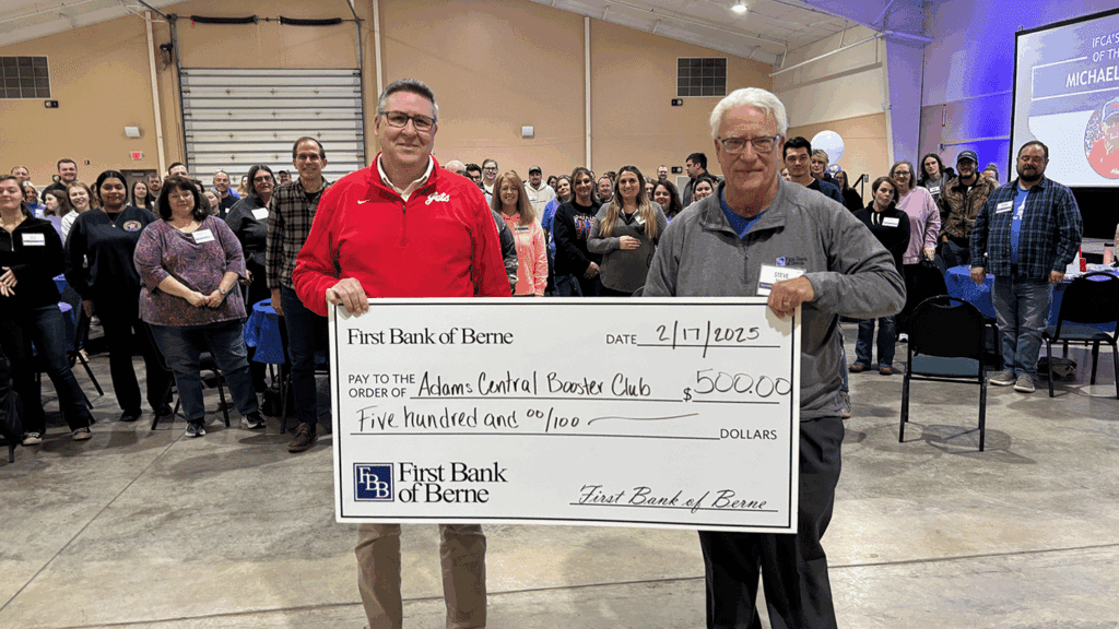 a photo showing two men holding a ceremonial large check representing a charitable donation in front of a large room of approximately 100 people