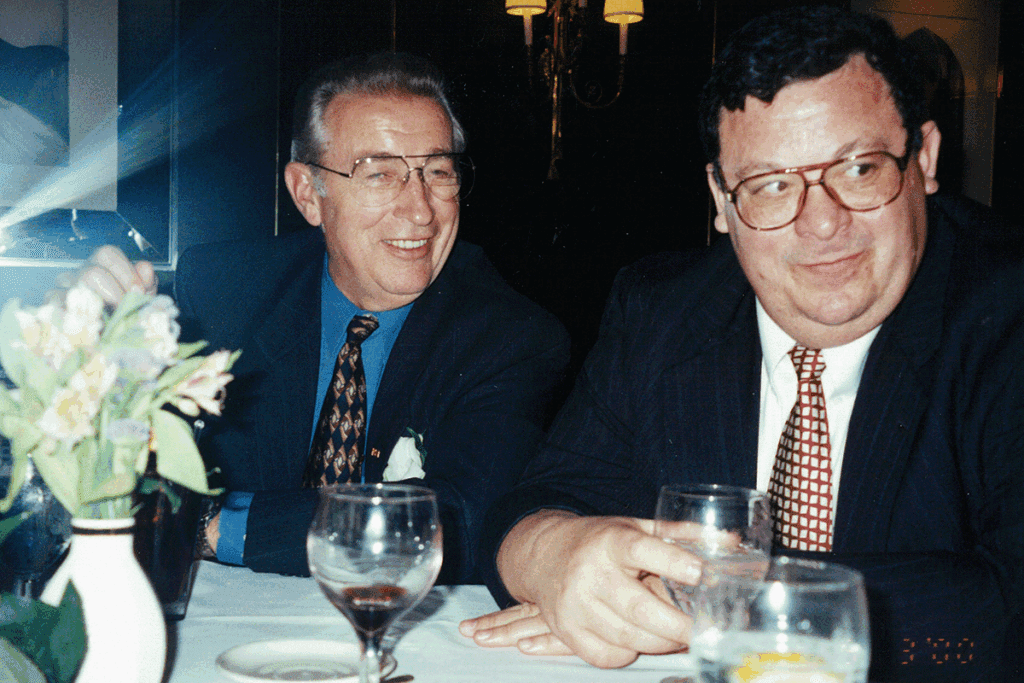 Two men wearing suits and ties are seen sitting at a banquet table, smiling as they look at something out of frame