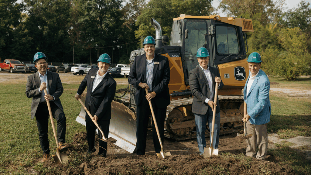 a photo showing five men wearing hard hats standing in front of an earth mover while holding shovels at a ceremonial groundbreaking