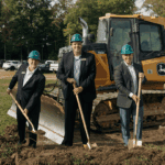 a photo showing five men wearing hard hats standing in front of an earth mover while holding shovels at a ceremonial groundbreaking