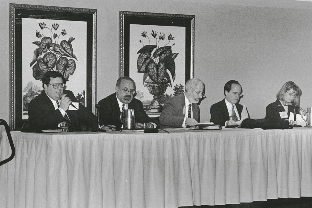 A black-and-white photo showing four men and a woman, all wearing professional clothing, sitting at a banquet table addressing an audience not seen in the photo