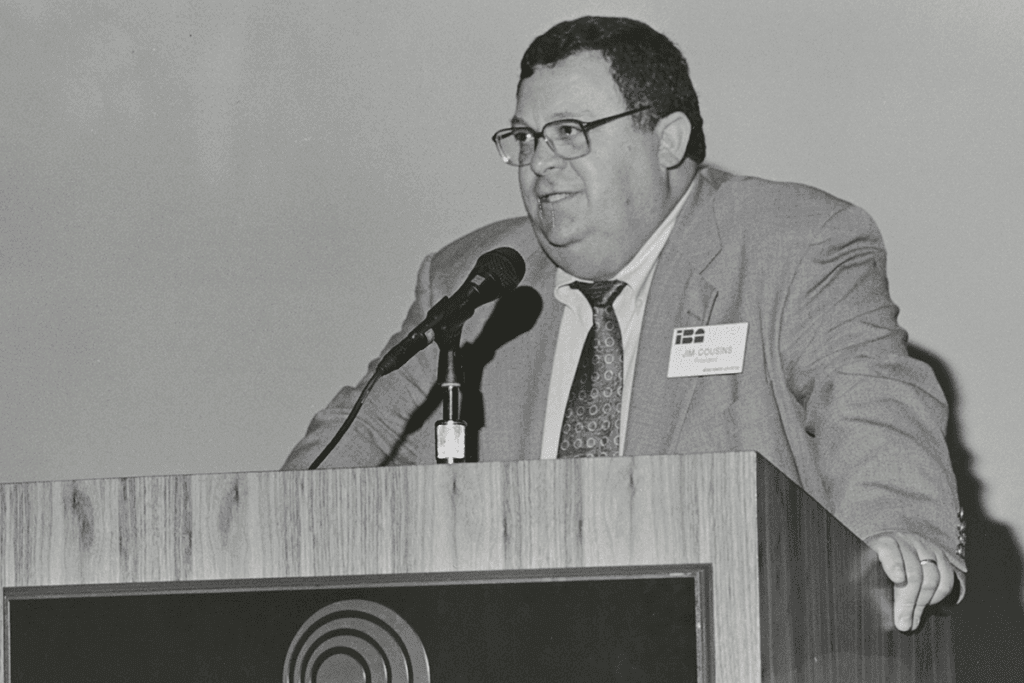 a man in a suit and tie addresses an audience not seen in the photo as he stands behind a podium