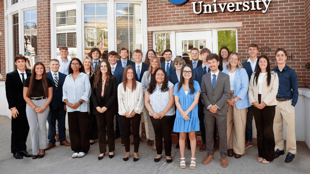 A photo showing 25 college students in business casual clothing standing in front of a brick building with the Lake City Bank logo