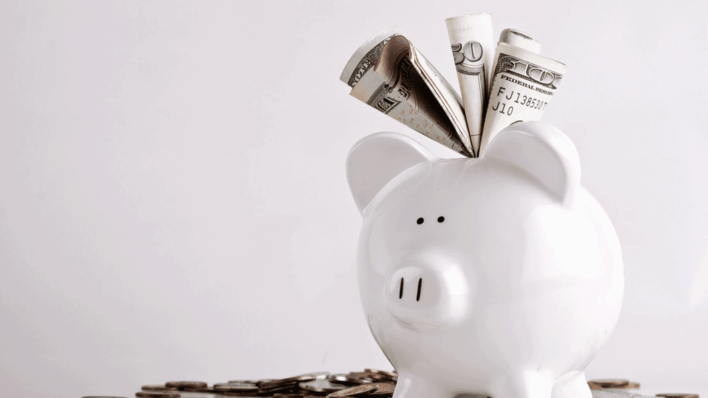 a picture showing a white piggy bank with dollar bills sticking out its top because it is so full, standing on a pile of coins