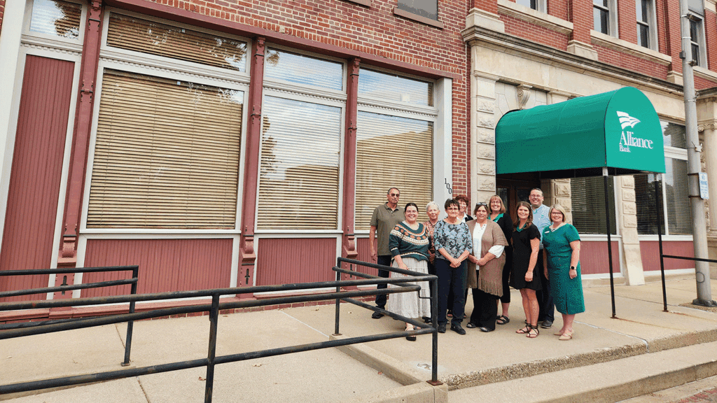 a group of 10 adults in business casual clothing stand in front of an historic brick building with a green awning covering a doorway behind them
