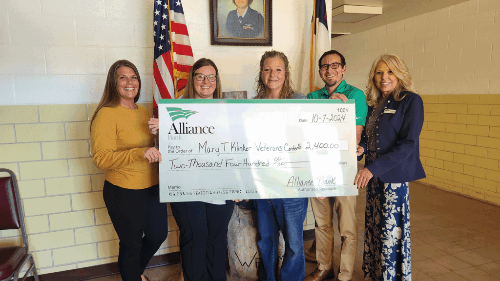 A group of five adults in casual clothing hold a large ceremonial check depicting a donation for $2,400, while standing inside a church building.