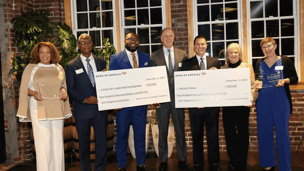 A line of 7 adults in professional clothing hold two ceremonial checks and related award trophies in front of a brick wall with windows.