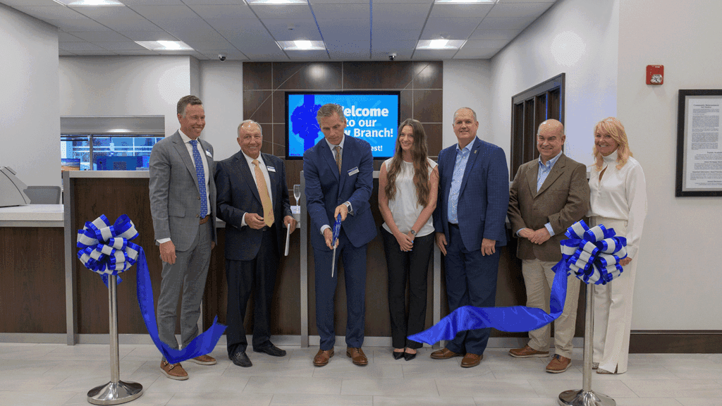 A line of seven people in professional clothing ceremonially cut a ribbon inside a modern bank lobby.