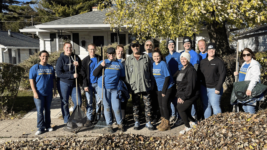 A line of 14 adults in outdoor work clothing stand in front of a modest home behind a large pile of leaves they have just raked out of the home's yard.