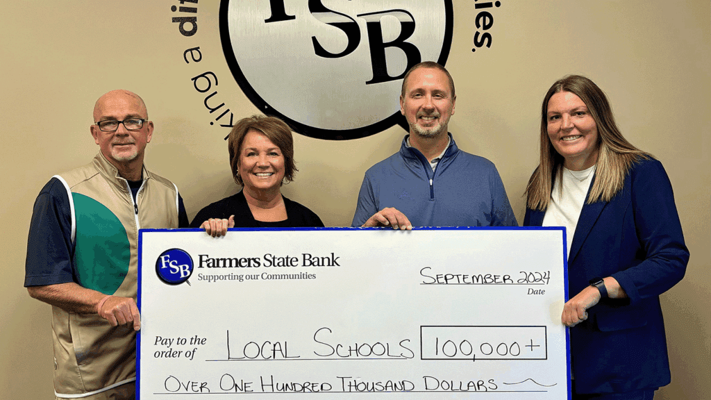 Four adults standing against a beige office wall hold a large ceremonial depicting a collective $100,000 donation to local schools.