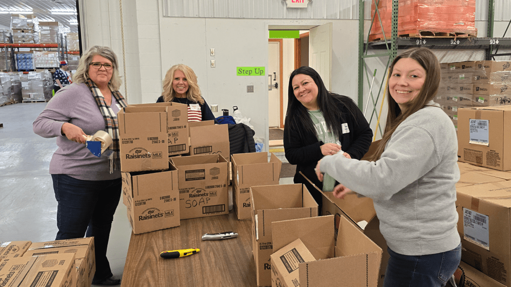 a picture showing four women standing in a warehouse in front of a pile of cardboard boxes they are building