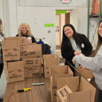 a picture showing four women standing in a warehouse in front of a pile of cardboard boxes they are building