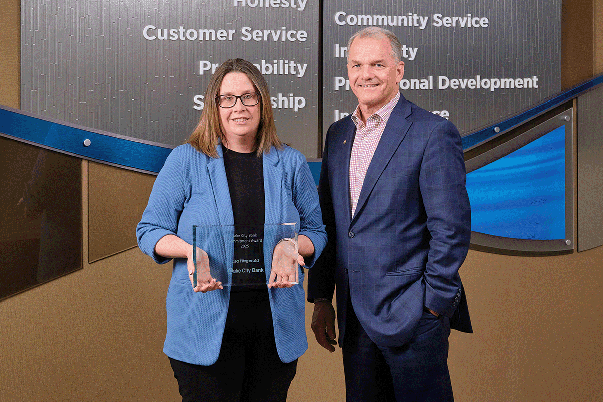 a photo showing a man and woman, both in professional clothing, as the woman in a light blue jacket holds in front of herself a glass award the man has just presented to her