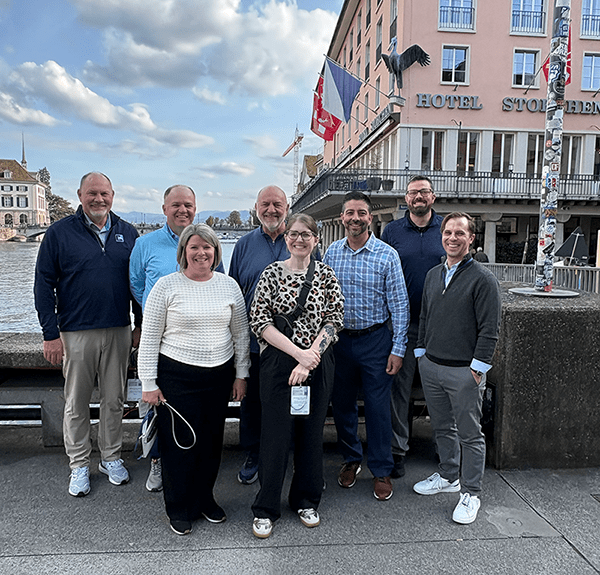 A photo showing eight people in business casual clothing standing in front of classic Swiss-style buildings