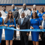 A photo showing 14 staff members in blue-themed professional clothing holding a blue ribbon to be cut at the unveiling of a new business, seen behind them