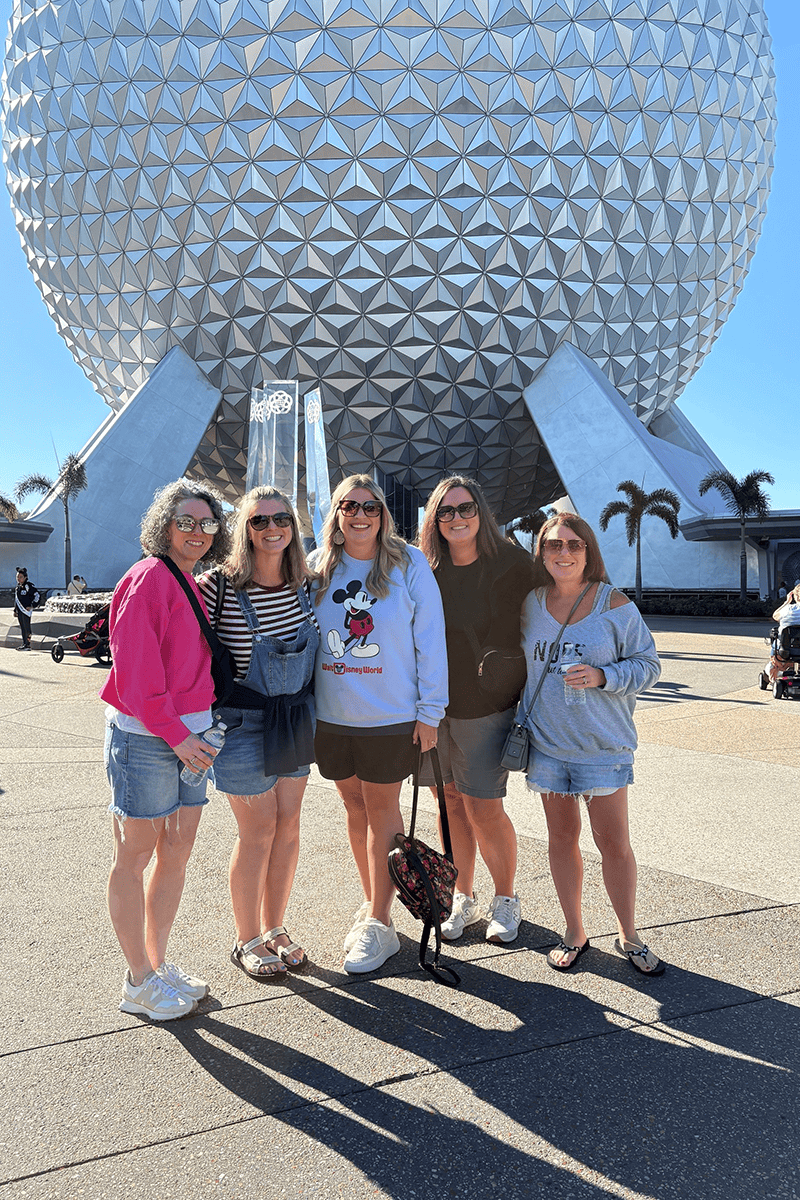 Five friends posing together in front of EPCOT's Spaceship Earth, wearing casual summer outfits and smiling at the camera.