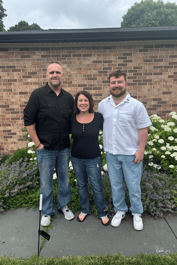 Three adults stand together outdoors in front of a brick building, smiling, with flowering bushes nearby.