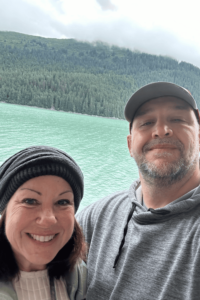 Smiling couple taking a selfie by a turquoise lake with a forested mountainside in the background.