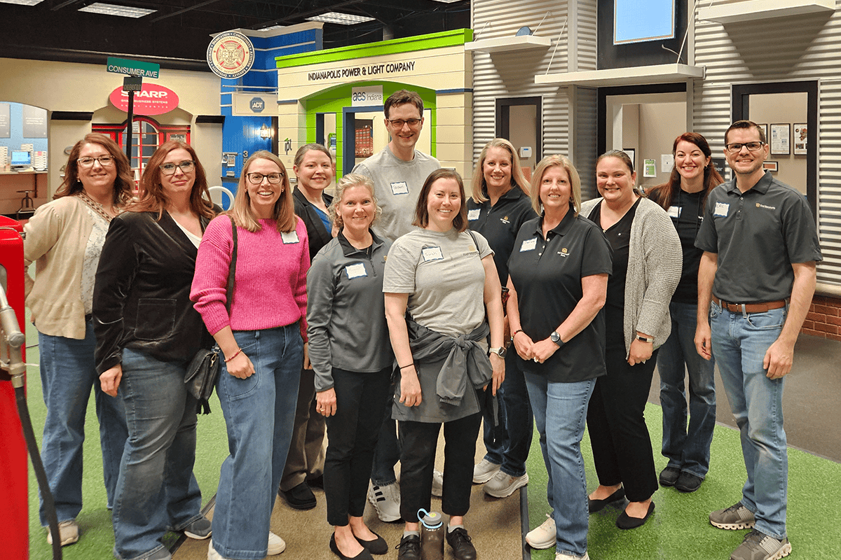 Group of smiling adults posing for a photo inside a museum-like exhibit or showroom, wearing name tags and casual attire.