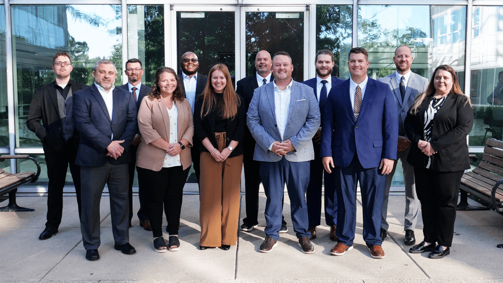 Group of twelve professionals in business attire posing outside a glass-front office building for a team photo.