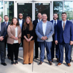 Group of twelve professionals in business attire posing outside a glass-front office building for a team photo.