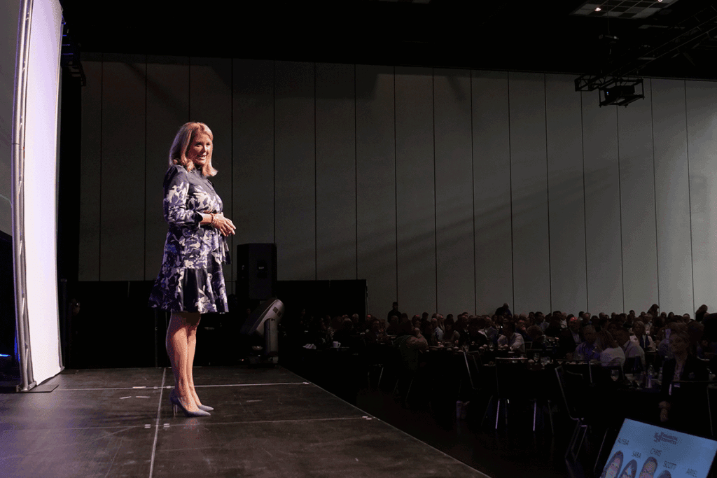 Woman in a blue floral dress stands on a stage and speaks to a seated conference audience in a dimly lit auditorium.