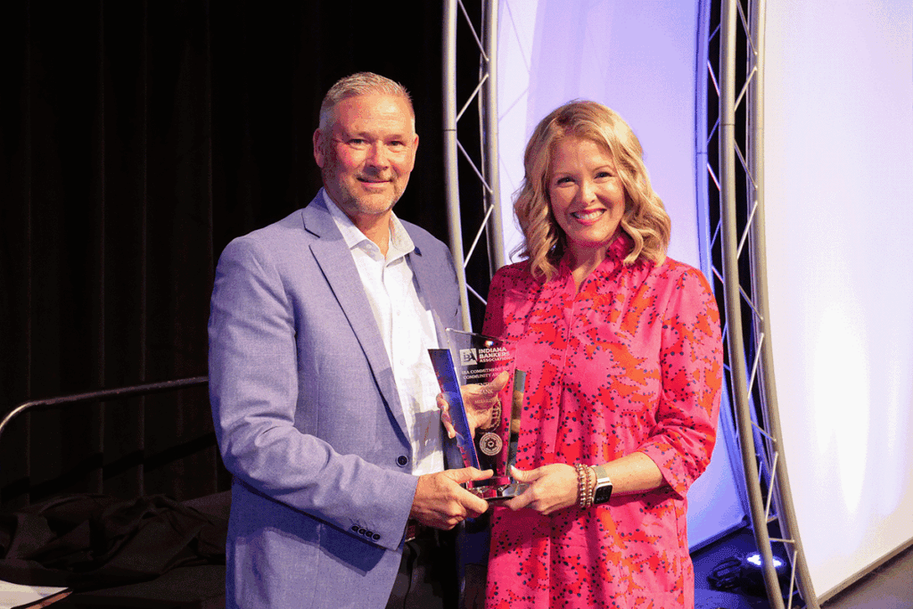 Man in a blue blazer presents a glass trophy to a smiling woman in a pink patterned dress on a stage.