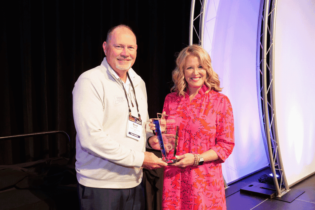 Man in a light sweater hands a glass award to a smiling woman in a pink patterned dress on stage, both wearing event lanyards.