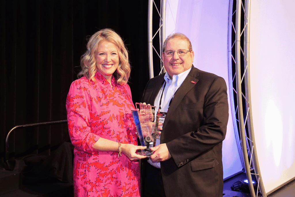 Smiling woman in a pink patterned dress and man in a suit hold a glass award on a stage.