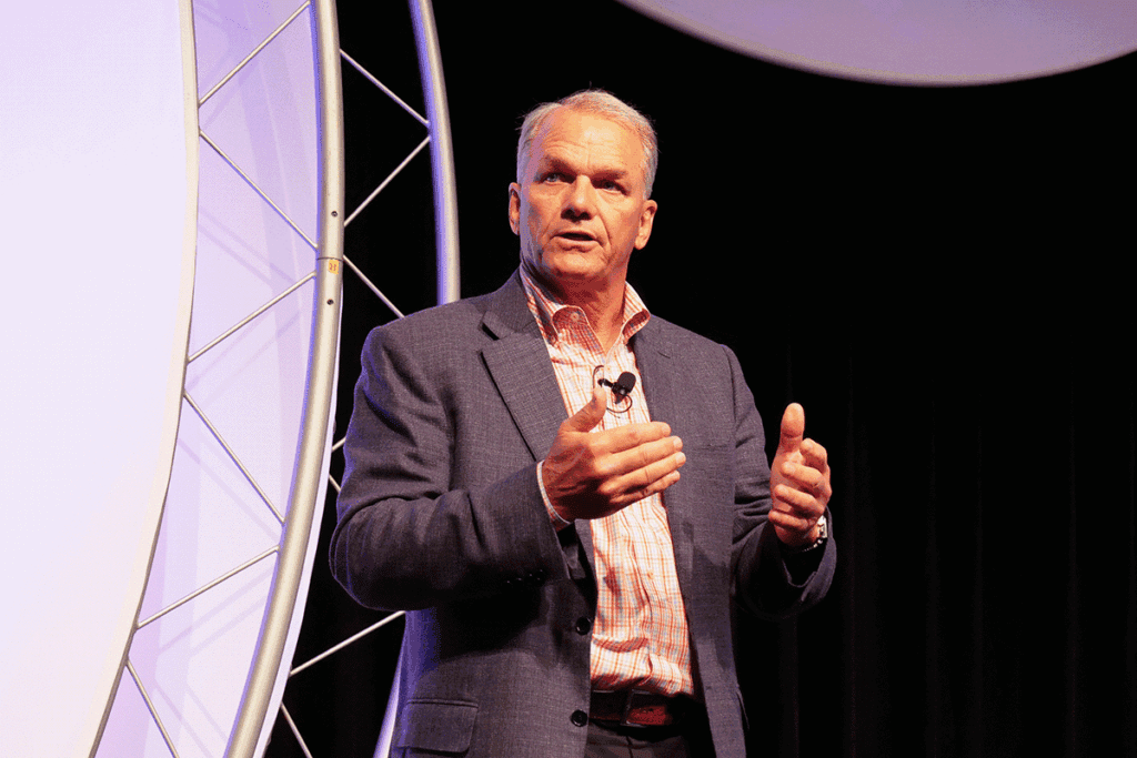 Male conference speaker in a gray blazer delivering a presentation on stage with a lapel mic