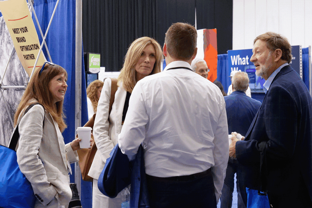Group of professionals conversing at a trade show booth, one woman in white jacket holding a coffee cup, others with blue tote bags nearby