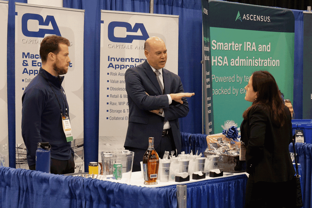 Two men in business attire at a trade show booth with large banners, talking to a woman attendee near a table with cups and a gift basket.