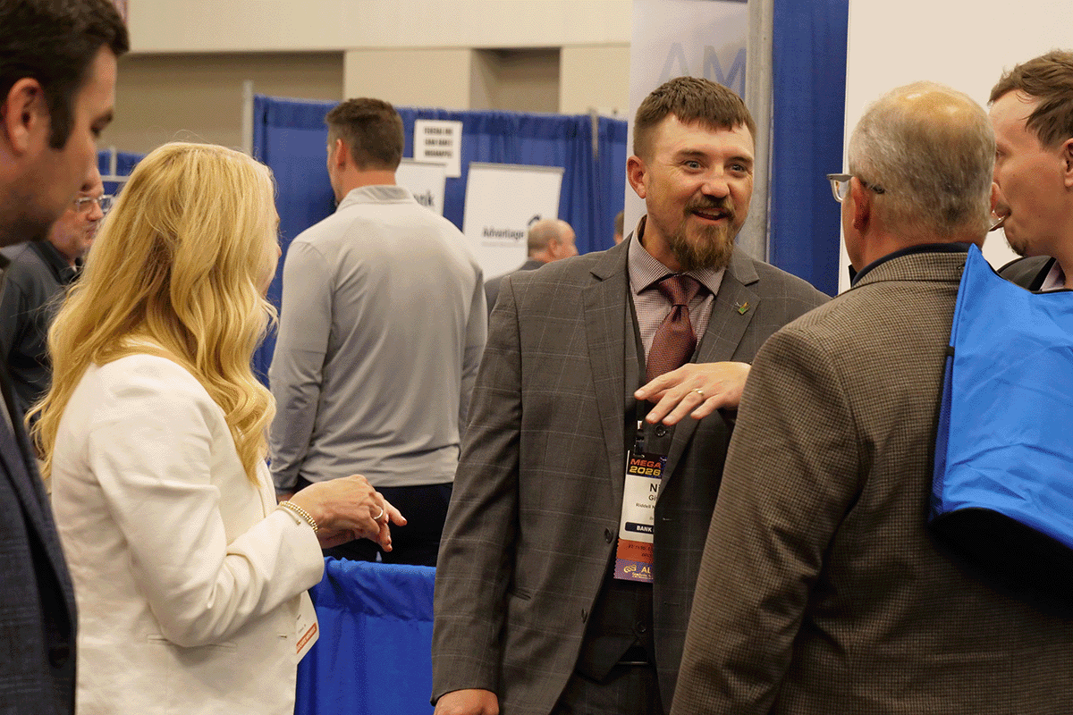 Two men in suits converse at a trade show, with a woman in a white jacket nearby, near blue booth curtains.