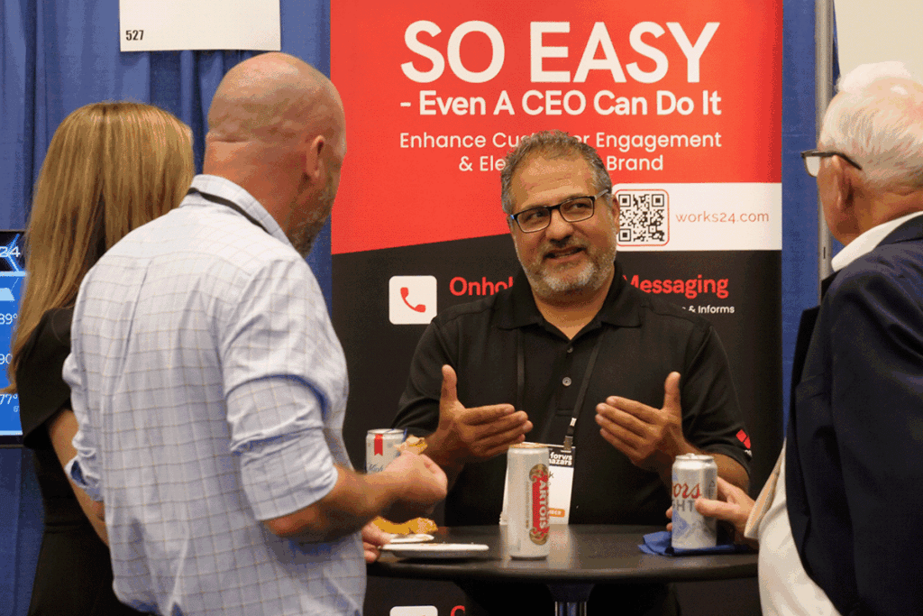 Man in black polo explaining something to two visitors at a trade show booth, with a red 'SO EASY' banner in the background and drinks on the table.