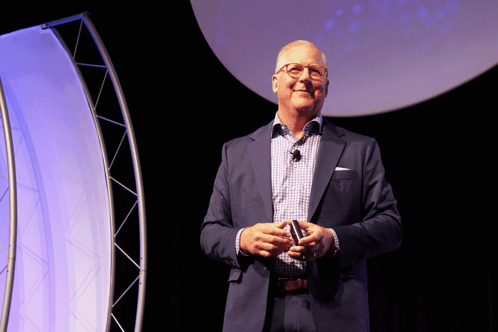 Man in a navy suit with a checkered shirt, smiling on stage with a clip-on mic, holding a small device; large circular purple backdrop behind him.