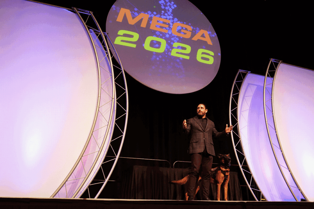 Man in a suit on stage with a dog beside him, under a large circular MEGA 2026 banner on stage lighting.