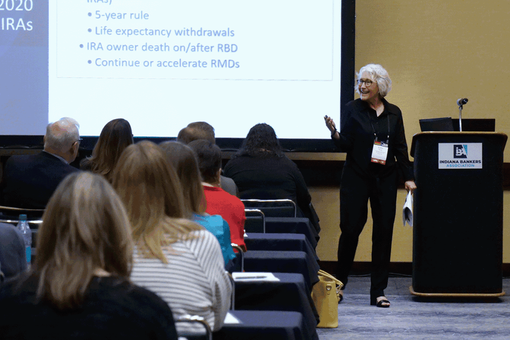 Presenter at a podium during an Indiana Bankers Association conference; slide with bullet points about IRAs is projected on a screen; audience seated in rows listening