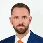 Professional headshot of a man with short brown hair and a beard, wearing a blue suit and orange tie against a white background.