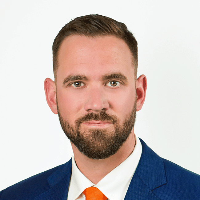 Professional headshot of a man with short brown hair and a beard, wearing a blue suit and orange tie against a white background.